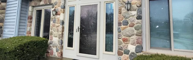 Front entry door with arched glass, side windows, stone exterior detail, and snow falling near the home