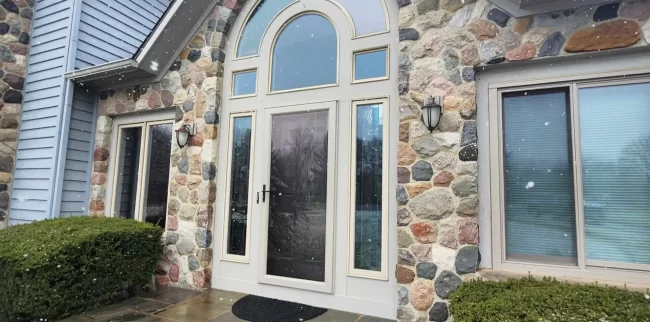 Front entry door with arched glass, side windows, stone exterior detail, and snow falling near the home