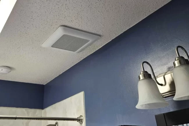 bathroom-exhaust-fan-replacement-aspect-ratio-960-640 Bathroom exhaust fan installed in a textured ceiling above a shower area with blue walls and vanity lighting