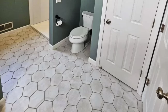 bathroom-hex-tile-flooring-update-aspect-ratio-960-640 Updated bathroom floor with light hexagon tile, gray-green walls, a white toilet, and shower entry
