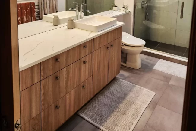 bathroom-vanity-update-aspect-ratio-960-640 Updated bathroom with wood vanity, white countertop, vessel sink, gray tile floor, and glass shower door.