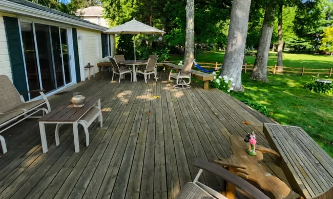Before view of older wood lakefront deck with patio furniture, weathered decking, and mature trees surrounding the backyard