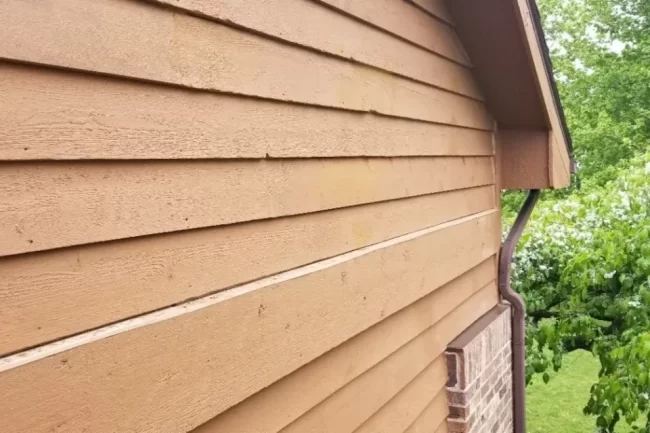 exterior-wood-siding-repair-1-aspect-ratio-960-640 Close-up of repaired exterior wood siding on a home with tan siding, brick detail, and surrounding greenery