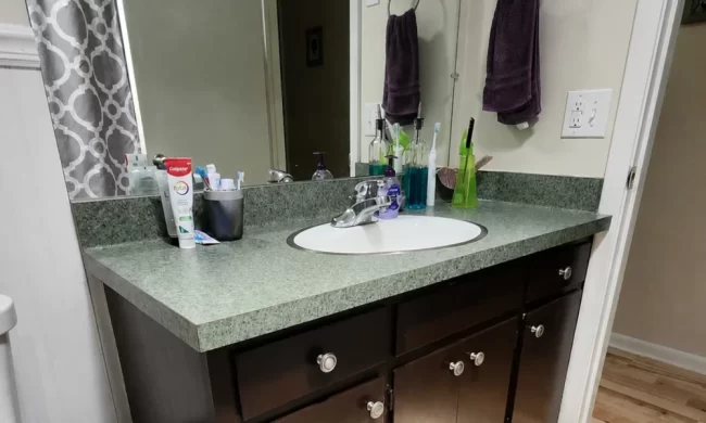 Before view of bathroom vanity with dark cabinet, green countertop, large frameless mirror, and older vanity lighting before replacement