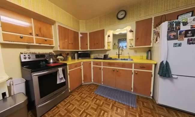 Before photo of the original kitchen with yellow countertops, dated cabinet fronts, patterned flooring, wallpaper border, white refrigerator, and older finishes before the kitchen update