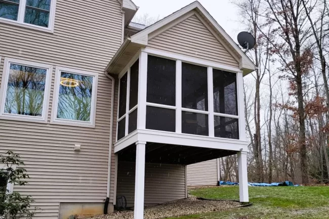 Elevated screened porch update with white support posts, dark screen panels, beige siding, and a wooded backyard