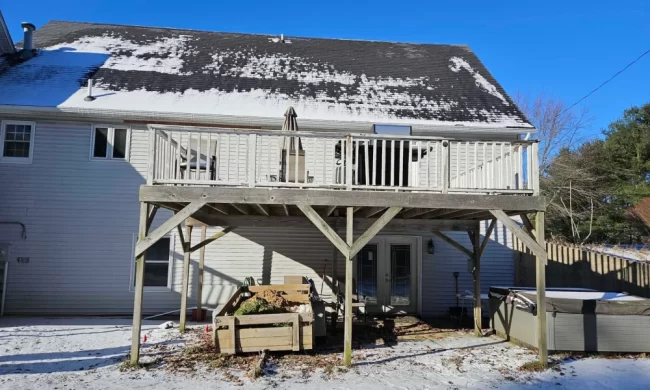 Before photo of an aging elevated backyard deck with weathered wood decking, white railings, exposed support posts, and snow-covered yard below