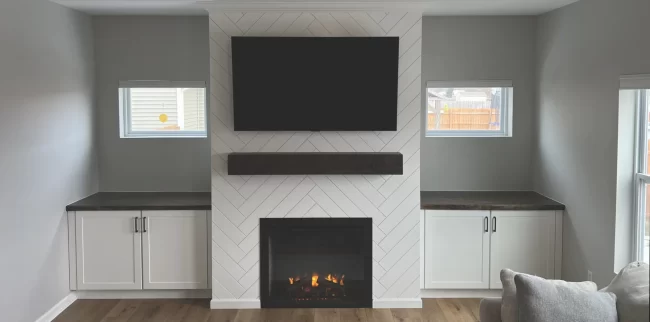 Living Room Fireplace and Built-In Cabinets After | Pennings Handyman After photo of a living room feature wall with a herringbone fireplace surround, dark wood mantel, mounted TV, and white built-in cabinets