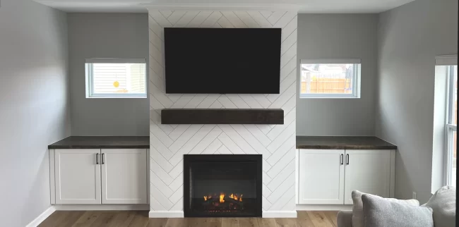 Living Room Fireplace and Built-In Cabinets After | Pennings Handyman After photo of a living room feature wall with a herringbone fireplace surround, dark wood mantel, mounted TV, and white built-in cabinets