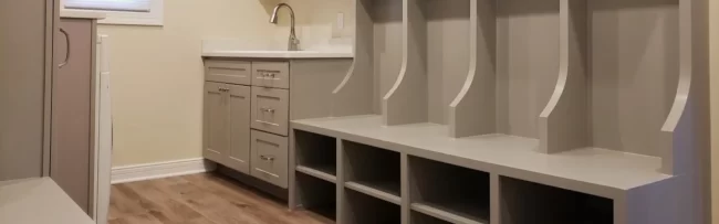 mudroom-cubbies-custom-carpentry-1-aspect-ratio-1920-600 Custom mudroom built-ins with storage cubbies, hooks, upper shelves, and cabinetry beside a small sink area