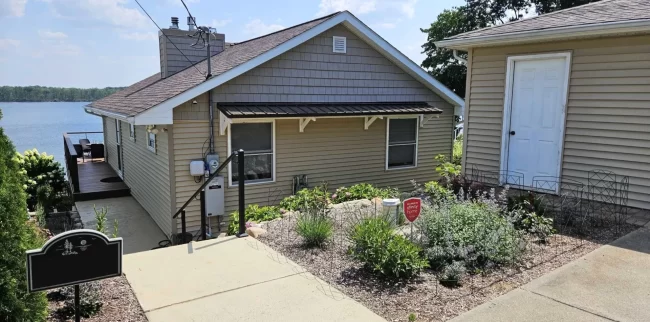 Lakefront home exterior with new handrail on stairs, metal awning, garden landscaping, and water view in the background