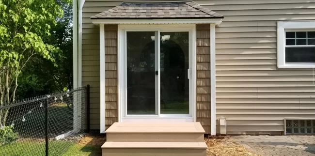 Sliding patio door with white trim, small entry steps, shingle awning, and tan home siding