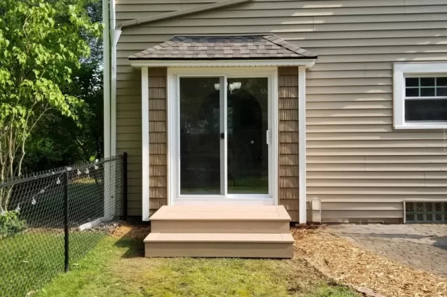 Sliding patio door with white trim, small entry steps, shingle awning, and tan home siding