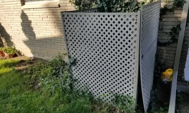Before photo of an older lattice waste container enclosure beside a brick exterior wall with overgrown grass and ivy
